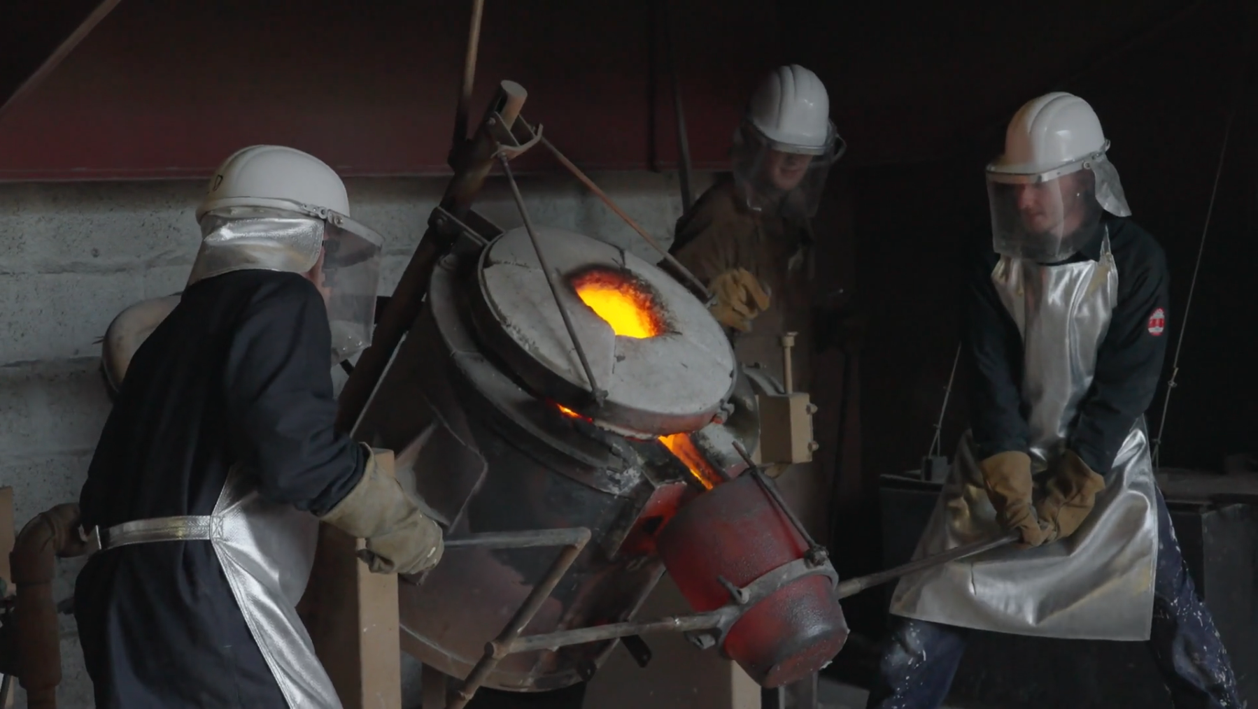 Three foundry workers wearing protective equipment tilt a crucible of glowing molten bronze during a pour